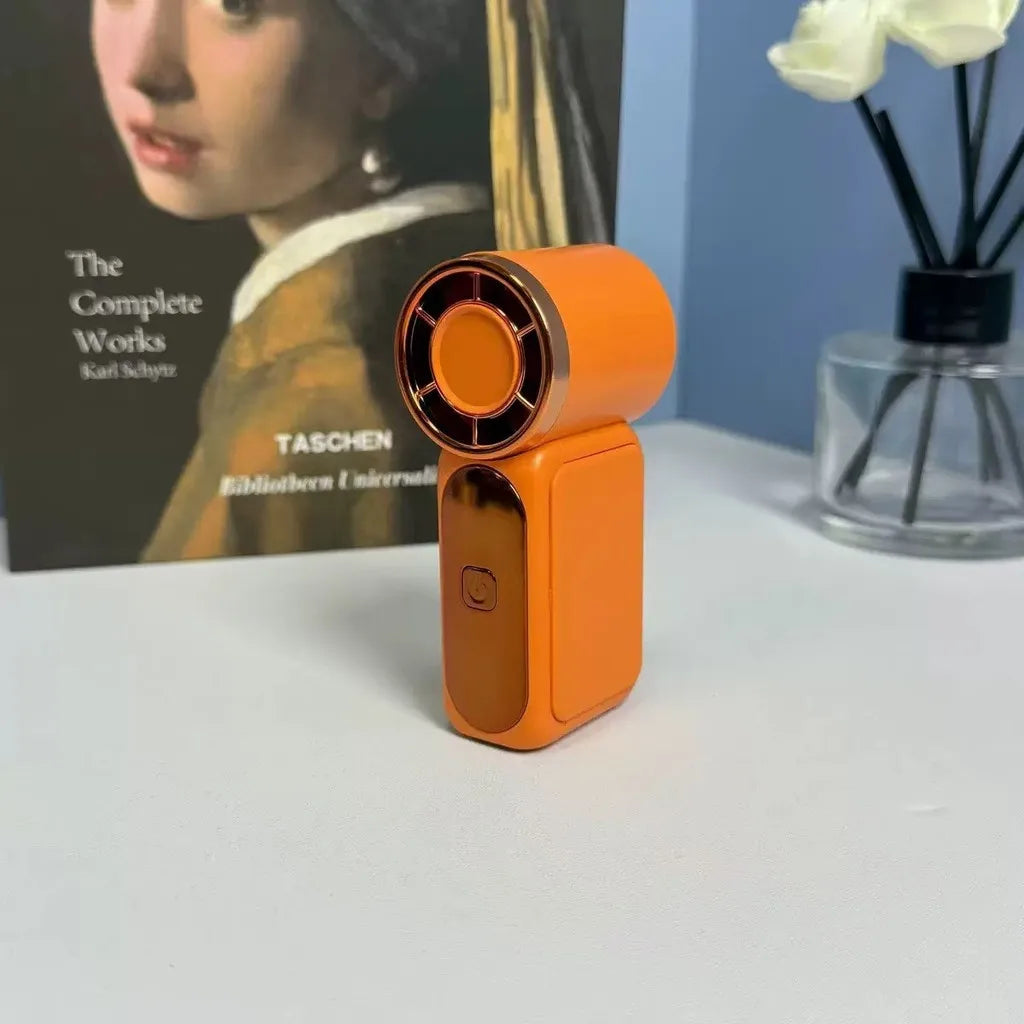 Orange portable mini fan with gold trim on desk next to book and reed diffuser – lifestyle shot